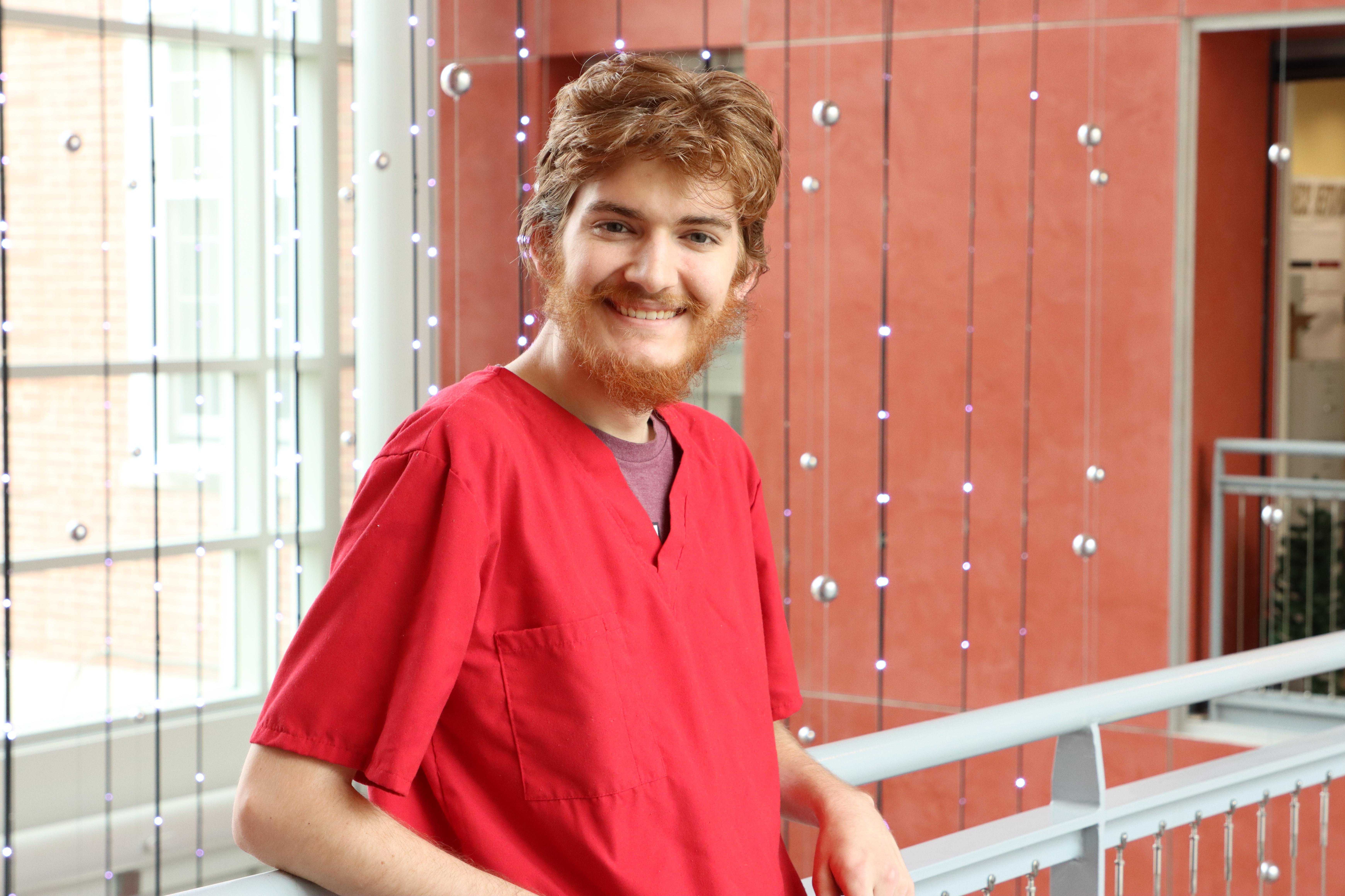 Jack, smiling and wearing a red scrub top with hanging lights behind him in the Toll Atrium. 